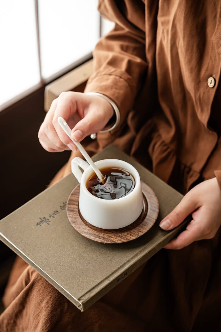 A person in warm linen holds a cup of coffee resting on a book, beside a sunlit window
